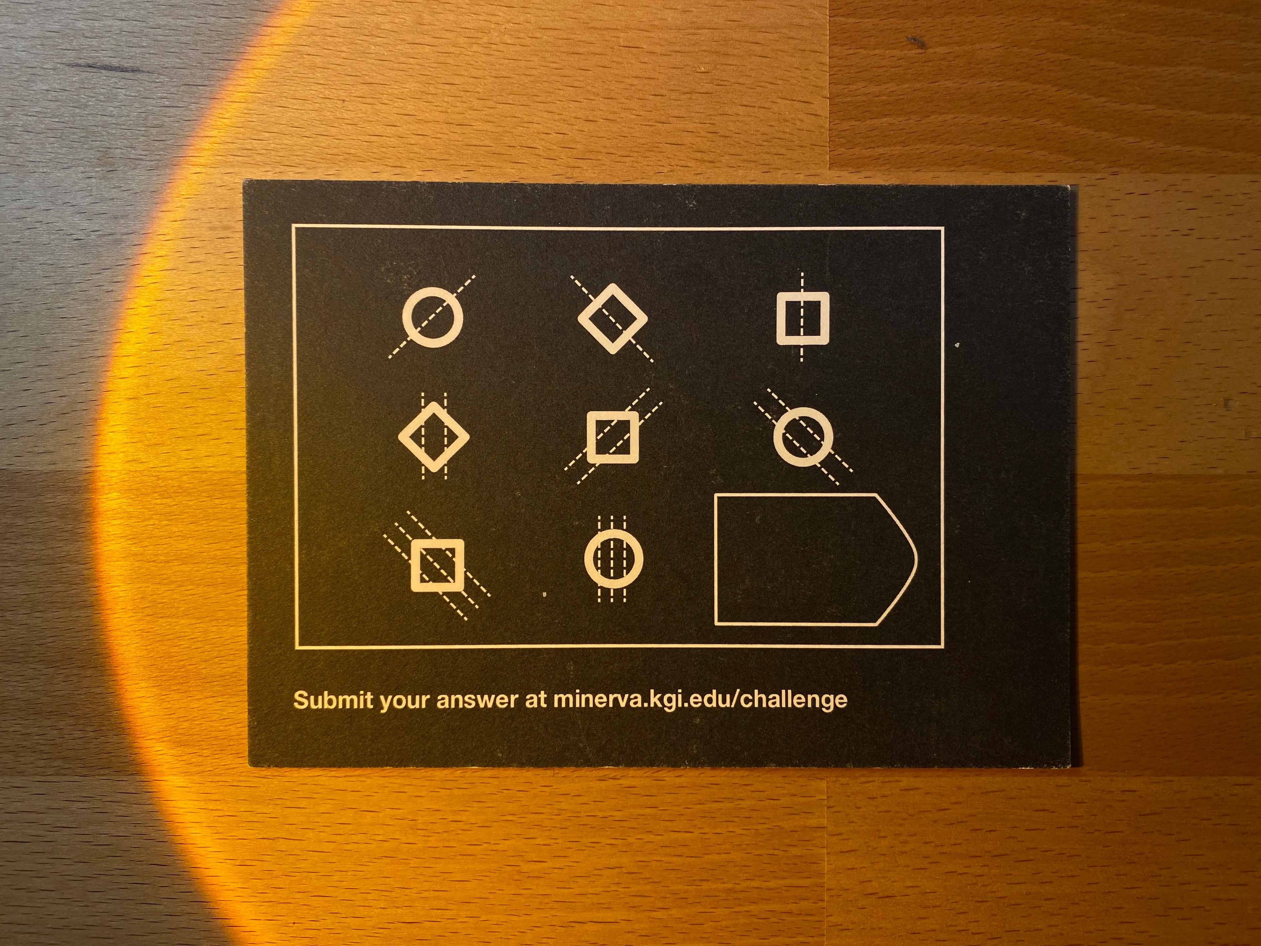 The postcard puzzle — back of the card with a 3x3 grid of shapes and the prompt 'Submit your answer at minerva.kgi.edu/challenge'